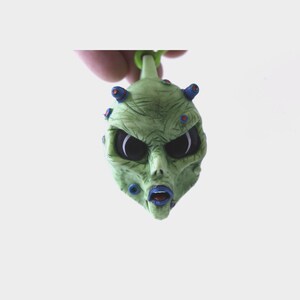 May include: Green alien head keychain with blue and red details. The alien has large black eyes and a wide open mouth with a blue tongue.