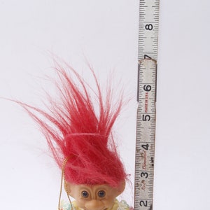 May include: A red-haired troll doll with a yellow and green dress. The doll is about 5 inches tall and is standing next to a ruler.