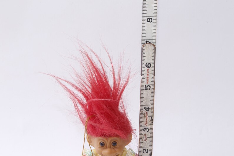 May include: A red-haired troll doll with a yellow and green dress. The doll is about 5 inches tall and is standing next to a ruler.