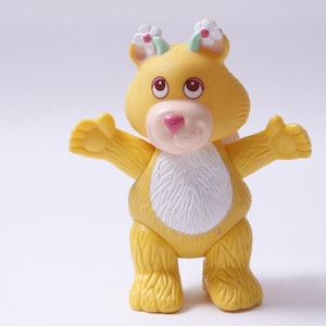 Könnte beinhalten: Gelbe Bärenfigur mit ausgestreckten Armen, einer rosa Nase und einem weißen Fleck auf der Brust. Der Bär hat kleine Blumenakzente auf dem Kopf und steht auf einer weißen Oberfläche. Ein Vintage-Spielzeug.