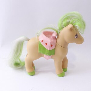 May include: A light brown toy pony with a green and pink saddle. The pony has a green and white mane and tail.