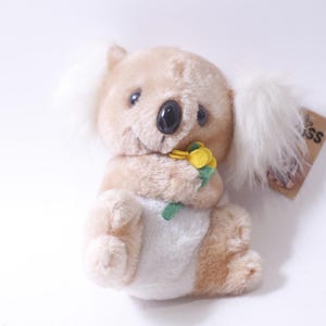 May include: A soft toy koala bear in shades of beige and white, with a black nose and a yellow flower. The koala has a tag attached. The toy is designed to look like a koala.