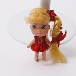 May include: A vintage doll with blonde hair styled in a side ponytail. The doll has blue eyes and is wearing a red dress. The doll is a miniature collectible.