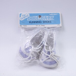 May include: A pair of doll baby running shoes in a clear plastic package. The shoes are white with light purple stripes and white laces. The package has a blue and white label that reads "DOLL BABY RUNNING SHOES". The shoes are 2.5 inches long.