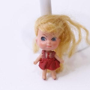 May include: A small vintage doll with blonde hair and blue eyes. The doll is wearing a red dress with a matching bodice. The doll is standing on a white surface.