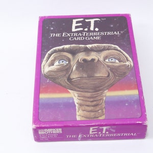 Puede incluir: Una caja morada para el juego de cartas E.T. El Extraterrestre. La caja presenta una imagen de primer plano de la cara de E.T. con una expresión amigable. El texto en la caja dice "E.T. El juego de cartas Extraterrestre".