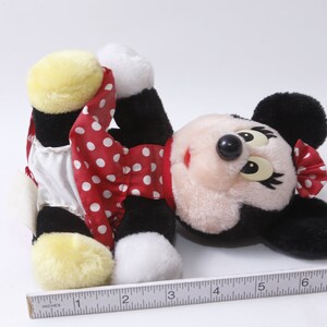 K&ouml;nnte beinhalten: Ein Pl&uuml;schtier von Minnie Mouse, die ein rotes Kleid mit wei&szlig;en Punkten und einen wei&szlig;en Unterrock tr&auml;gt. Sie hat schwarze Ohren und eine schwarze Nase. Ihre Augen sind geschlossen und sie liegt auf dem R&uuml;cken.