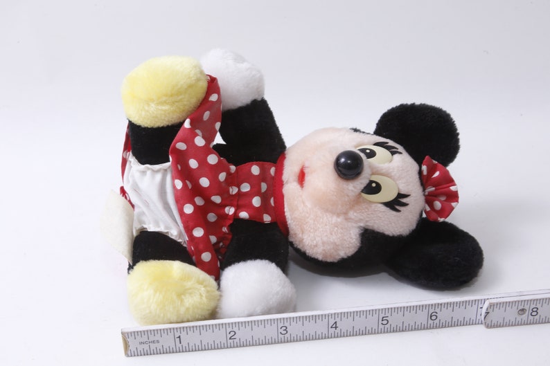K&ouml;nnte beinhalten: Ein Pl&uuml;schtier von Minnie Mouse, die ein rotes Kleid mit wei&szlig;en Punkten und einen wei&szlig;en Unterrock tr&auml;gt. Sie hat schwarze Ohren und eine schwarze Nase. Ihre Augen sind geschlossen und sie liegt auf dem R&uuml;cken.