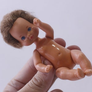 May include: A small plastic baby doll with brown hair and blue eyes. The doll is nude and has a painted belly button.