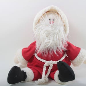 May include: A plush Santa Claus doll with a white beard and red suit. The doll is sitting with its legs crossed and has a white rope belt.
