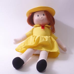 May include: A handmade doll with red hair, wearing a yellow dress and matching hat. The doll has a red bow and black shoes. The doll is a soft, plush toy.
