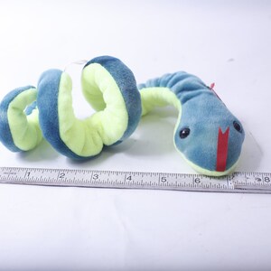Pode incluir: Um brinquedo de cobra de pel&uacute;cia azul e verde com uma boca vermelha. A cobra est&aacute; enrolada e mede cerca de 15 cm de comprimento.
