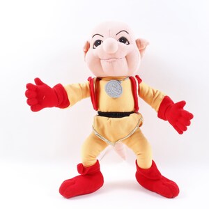 May include: A plush toy of a pink-skinned character with large ears, wearing a yellow jumpsuit with a silver medal around its neck. The character has a large nose and a mischievous expression.