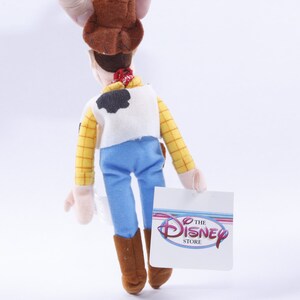 Może przedstawiać: Pluszowa zabawka Woody'ego z Toy Story. Ma na sobie sw&oacute;j charakterystyczny kowbojski kapelusz, kamizelkę i dżinsy. Zabawka trzyma metkę z napisem "The Disney Store".