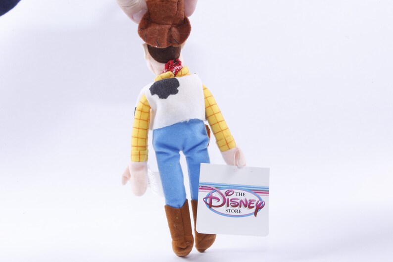 Może przedstawiać: Pluszowa zabawka Woody'ego z Toy Story. Ma na sobie sw&oacute;j charakterystyczny kowbojski kapelusz, kamizelkę i dżinsy. Zabawka trzyma metkę z napisem "The Disney Store".