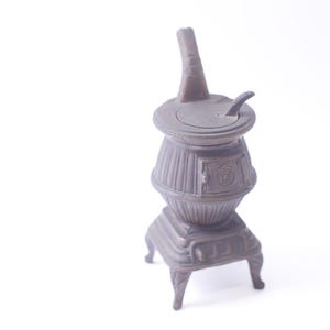 May include: A vintage-style metal stove pencil sharpener with a black finish and a hinged lid. The sharpener has a decorative design and a small handle on the lid.