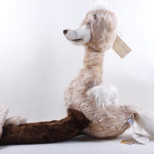 May include: A plush stuffed animal featuring a long, slender neck and a head with light tan fur. The body has a mix of light tan and white fur, while the extended lower legs are covered in dark brown fur. A small paper tag is attached to the neck.