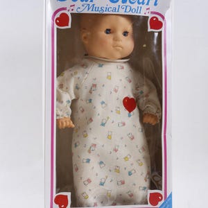 Könnte beinhalten: Eine Vintage-Musikpuppe "Dear Heart" in ihrer Originalverpackung. Die Puppe trägt einen weißen Strampler mit einem Muster aus kleinen Bären und Herzen. Ein großes rotes Herz befindet sich auf der Brust der Puppe. Auf der Box steht "Die weltweit erste elektronische Chip-Musikpuppe."