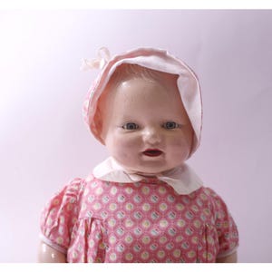Puede incluir: Muñeca vintage con rostro pintado, ojos azules y labios rojos. La muñeca viste un vestido rosa con un estampado geométrico y un gorro a juego con ribete blanco. La muñeca tiene un cuello blanco.