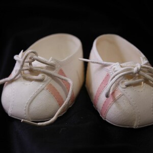 May include: A pair of white baby trainers with pink stripes and white laces. The shoes are made of a soft material and are designed for infants. The trainers are set against a black background.