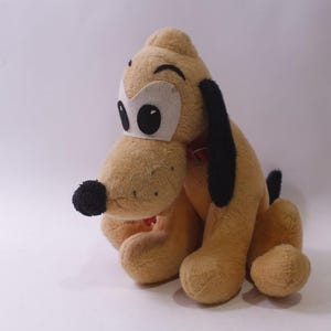 Peut inclure: Peluche représentant un chien de dessin animé, avec une fourrure beige, des oreilles noires et un nez noir. Le chien a de grands yeux ronds et un collier rouge. Le jouet est posé sur une surface blanche.