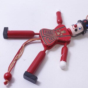 May include: A vintage wooden pull-string toy soldier with a red body, black accents, and a painted face. The toy has jointed limbs and a decorative bow tie. The soldier's hat is black with white and blue dots.