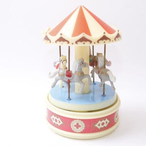 May include: A miniature carousel toy with a red and cream striped canopy. The carousel features several toy horses in various colors, mounted on a light blue base. The base is decorated with a red band with white floral designs.