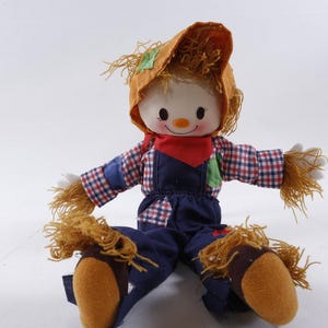 May include: A handmade scarecrow doll with a smiling face, wearing a straw hat, red bandana, and blue overalls. The doll has straw hair, arms, and legs, and is sitting with its legs outstretched. A charming, whimsical decorative item.