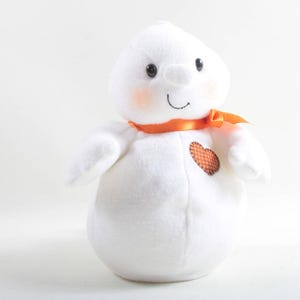 May include: A white snowman plush toy with black eyes, a smiling mouth, and rosy cheeks. It has an orange ribbon around its neck and a heart-shaped patch on its chest. The snowman is a festive decoration.