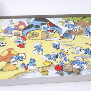 K&ouml;nnte beinhalten: Ein Spielbrett zum Thema Schlumpf mit einem Pilzhaus, einem Fluss und verschiedenen Schlumpf-Charakteren. Die Figuren sind blau mit wei&szlig;en H&uuml;ten und in verschiedenen Posen dargestellt. Das Spielbrett ist aus Pappe und in gutem Zustand.