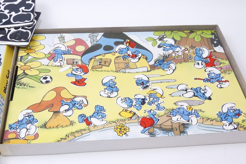K&ouml;nnte beinhalten: Ein Spielbrett zum Thema Schlumpf mit einem Pilzhaus, einem Fluss und verschiedenen Schlumpf-Charakteren. Die Figuren sind blau mit wei&szlig;en H&uuml;ten und in verschiedenen Posen dargestellt. Das Spielbrett ist aus Pappe und in gutem Zustand.