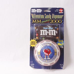 Op de afbeelding: M&M's Millennium Candy Dispenser, een verzamelobject in beperkte oplage. De verpakking heeft een blauwe achtergrond met de tekst "MM means 2000". De dispenser heeft een rond ontwerp met de tekst "The Official Candy of the New Millennium".