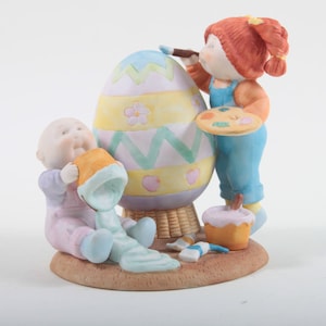 Può includere: Un set di figurine in porcellana che raffigurano due bambini che dipingono un uovo di Pasqua. La ragazza ha i capelli rossi e tiene un pennello e una tavolozza. Il ragazzo tiene un secchio di vernice. L'uovo è decorato con strisce colorate e fiori.