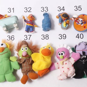 May include: A collection of Sesame Street character figurines and plush toys. The figurines include Ernie, Cookie Monster, and Bert. The plush toys include Elmo, a green character with orange hair, and a yellow duck.