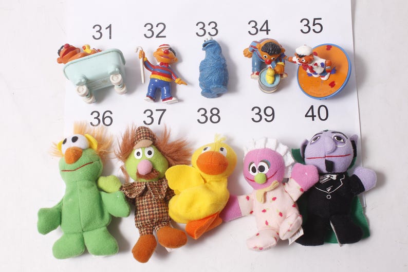 May include: A collection of Sesame Street character figurines and plush toys. The figurines include Ernie, Cookie Monster, and Bert. The plush toys include Elmo, a green character with orange hair, and a yellow duck.