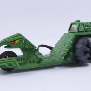 May include: Green plastic toy vehicle with a bird-like head and a large wheel in the back. The vehicle has a detailed design with a textured surface.