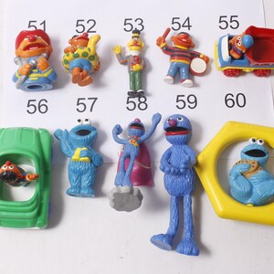 May include: A collection of Sesame Street character figurines, including Elmo, Cookie Monster, and Bert and Ernie. The toys are brightly colored and made of plastic. The figures are numbered 51 through 60.