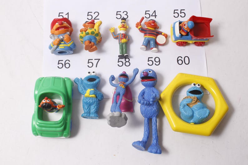 May include: A collection of Sesame Street character figurines, including Elmo, Cookie Monster, and Bert and Ernie. The toys are brightly colored and made of plastic. The figures are numbered 51 through 60.