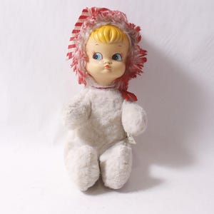 May include: A vintage doll with a yellow plastic face, blue eyes, and a red and white striped bonnet. The doll's body is made of white, fluffy material. It is sitting upright.