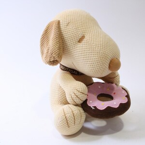 May include: A plush toy dog, beige with brown accents, holding a pink frosted donut with sprinkles. The dog has a collar with the text "UNIVERSAL STUDIOS". The toy is on a white background.