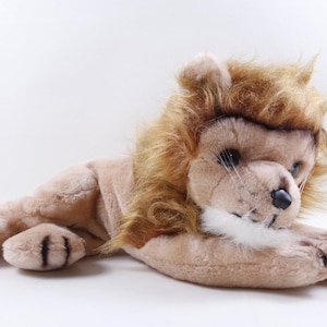 May include: A plush lion toy in a relaxed pose. The toy has a tan body, a brown mane, and a white muzzle. The lion's paws have black stripes, and it has a black nose and whiskers. The toy is lying down on a white surface.