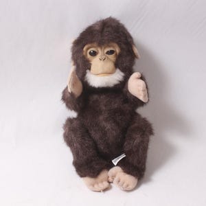Op de afbeelding: Een pluchen aap met bruine vacht, bruine handen, voeten en oren en een witte vlek rond de mond. Het speelgoed heeft een realistisch gezicht met grote ogen en zit rechtop.