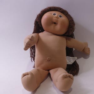 Op de afbeelding: Een vintage Cabbage Patch Kid-pop met een zacht, bruin lichaam en een rond, bruin gezicht. De pop heeft bruin garen haar en is ongekleed. De armen en benen van de pop zijn uitgestrekt en ze heeft een kleine navel.