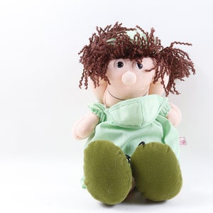 May include: A plush doll wearing a green surgical gown and a brown wig. The doll has a large nose and brown eyes.