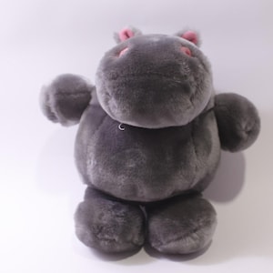 Op de afbeelding: Een grijze pluche nijlpaard met roze accenten op de oren en snuit. Het knuffeldier heeft een zachte, harige textuur en staat rechtop. Het nijlpaard heeft kleine, ronde armen en benen. Kinderspeelgoed.