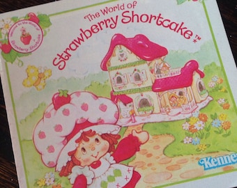 May include: A vintage children's book cover featuring Strawberry Shortcake, a cartoon character with a red dress and a strawberry hat, standing in front of her house. The book title is "The World of Strawberry Shortcake" with the trademark symbol.