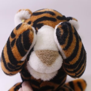 May include: A plush tiger toy with orange and black stripes, white accents, and a tan nose. The tiger's ears are positioned over its eyes, creating a playful, hiding effect. The toy is soft and cuddly, perfect for children.