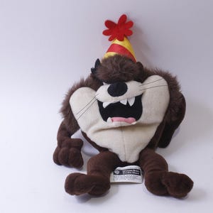 Könnte beinhalten: Ein braunes und weißes Plüschtier des Zeichentrickcharakters Taz aus Looney Tunes. Taz trägt einen roten und gelben Partyhut mit einer roten Blume oben drauf. Das Spielzeug sitzt auf einer weißen Oberfläche.