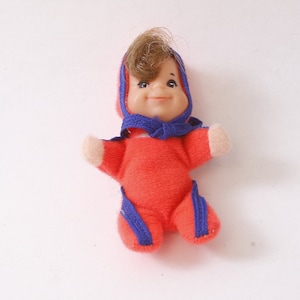 Peut inclure: Une petite poupée en peluche rouge avec une tenue à capuche bleue et des cheveux bruns. La poupée a un visage souriant et porte un nœud bleu.