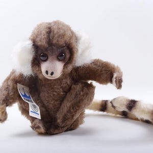 May include: A plush monkey toy with brown fur, white ear tufts, and a striped tail. The toy has a tag attached. The monkey is sitting on a white surface.
